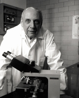 Robert Krasner in a lab coat stands behind a microscope, facing the camera, with a shelf and a brick wall in the background.