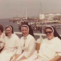 Three sisters sitting on a boat in a body of water. There are piers and buildings in the background. People are sitting on either side of the sisters.