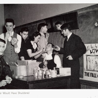 A classroom scene where several people are gathered around a seated individual having makeup applied, with a cluttered table and a poster in the background reading "Will he be a LOVER or THE FALL GUY."