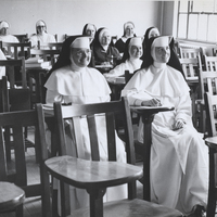 Several sisters in habits are sitting in classroom desks looking at the front of the room.
