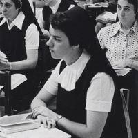 A nun sits at a desk in a classroom with a book in front of her. There are two other nuns sitting behind her.