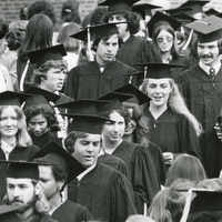 Several students wearing graduation regalia walk down an aisle outdoors as audience members stand on either side of the aisle.