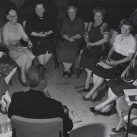 Several women sit in chairs organized into a semi circle in front of a man wearing a clerical suit.