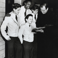A group of four young boys in school uniforms standing with a reverend. The boys are wearing light-colored blazers, dark ties, and dark pants. The adult man is dressed in a clerical suit and is holding a book or document that the group appears to be looking at together.