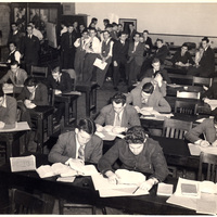 A crowded classroom with students in formal attire seated at desks, reading or writing, suggesting an exam or study session.