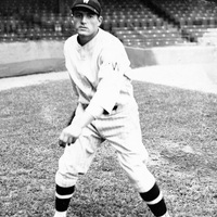 Eddie Wineapple in a pitching stance on a field, wearing a baseball uniform with striped socks and a cap marked with 'W,' with empty stadium seats in the background.
