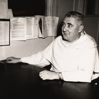 Rev. Vincent Dore in a light-colored robe seated at a desk, with documents pinned to the wall behind them in an office setting.