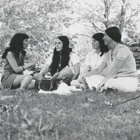 Four women sit on the grass outside. One woman is holding a microphone, interviewing the other three.
