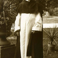 Father James Meagher standing outdoors in a garden, wearing a long white robe with a dark cloak, surrounded by potted plants and trees.