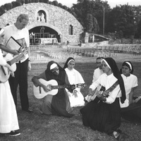 A group of nuns and a man gathered outdoors, playing guitars. In the background, there is an open-air stone structure with an archway and seating area. The nuns are dressed in traditional habits, and some are seated on the grass while others stand.