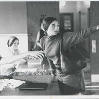 Black and white photograph showing three people in a room. One person is seated at a table with a box of files, reaching out towards another person who is standing and pointing to the right. The third person is partially visible in the background.