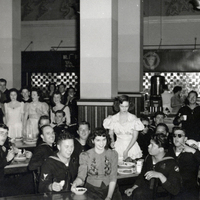 A group of people in a dining area. Some people are seated at tables eating, while others are standing. The setting appears to be a cafeteria or mess hall with several tables and chairs. In the background, there is a counter with food service equipment and tiled walls.