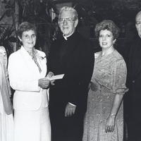 Three women and two men pose for a photograph. One woman is handing one of the men a check. The men are wearing clerical suits.