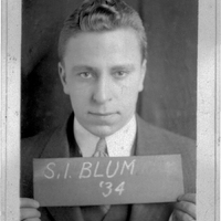 Photo of Samuel Blum in a suit and tie holding a sign that reads "S.I. BLUM '34," looking directly at the camera.