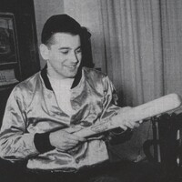 Aaron Slom seated indoors wearing a shiny jacket, holding a baseball bat and looking at it, with a framed picture and curtains visible in the background.