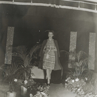 A woman wearing a dress and coat walks down a run way lined with plants.
