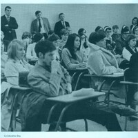 A classroom filled with students most of whom are seated at desks, some taking notes while others listen attentively. The image is labeled 'Co-Education Day' at the bottom left corner.