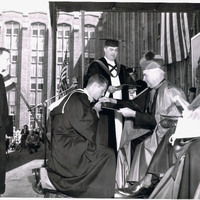 An outdoor formal ceremony with a person in academic regalia kneeling before a seated figure in ceremonial robes, receiving a document, with others in academic attire and American flags in the background.