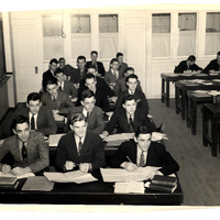 A classroom with students in formal attire seated at desks, reading or writing, with large windows and tables in the background.
