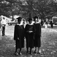 Three women standing on grass outside wearing graduation regalia. One graduate is holding a degree. There are several people in the background and a parked car.