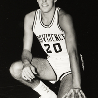 Stuart Kerzner in a PC jersey with the number 20 kneels on one knee, holding a basketball in one hand against a dark background.