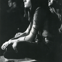 Two women sit on the floor of a dark room looking in one direction. There are some people also sitting on the floor in the background.