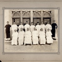 Fourteen clergy members posed in front of a building with Gothic arched windows, with seven seated in white robes and seven standing behind.