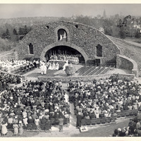 A large outdoor religious ceremony at a stone grotto, with clergy gathered around an altar and a seated crowd in attendance, set against the Providence cityscape.