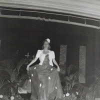 A woman wearing a long dress with a white jacket walks down the runway with plants along the side.