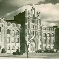 A Gothic-style building with arched windows and ornate stonework, vintage cars parked in front, and trees in the foreground.