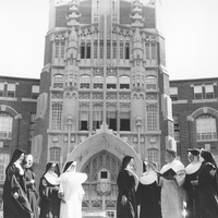 A group of nuns and a priest stand in front of a large, ornate building with Gothic architectural elements. The nuns are dressed in traditional habits, and the priest is wearing clerical robes.
