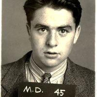 Morton Hoffman in a suit jacket, striped shirt, and tie holding a sign that reads "M.D. 45 HOFFMAN," against a plain background.