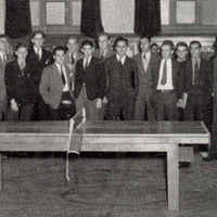 Two people playing table tennis in a large indoor room, with a group of formally dressed spectators watching closely from behind.
