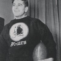 Aaron Slom standing indoors holding a football, wearing a dark sweatshirt with the word "Friars" and an image of a friar on the front, with curtains in the background.