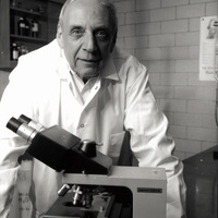 Robert Krasner in a lab coat stands behind a microscope, facing the camera, with a shelf and a brick wall in the background.