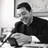 Rev. William Haas in clerical attire seated at a desk, smiling while looking down at an object held in both hands.